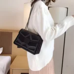 Sac à main noir matelassé avec chaîne cloutée porté sur l'épaule par une femme avec un pull blanc dans une chambre d'hôtel. On distingue la longueur de la chaîne du sac qui permet de le porte facilement à l'épaule sans aucune gène.