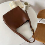Sac à main marron à bandoulière réglable en simili cuir, posé sur une table blanche avec un livre et une tasse à café
